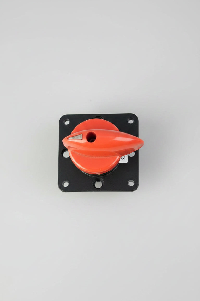 An industrial toggle switch with a bright orange-red handle mounted on a black square base, photographed from above on a textured red surface.